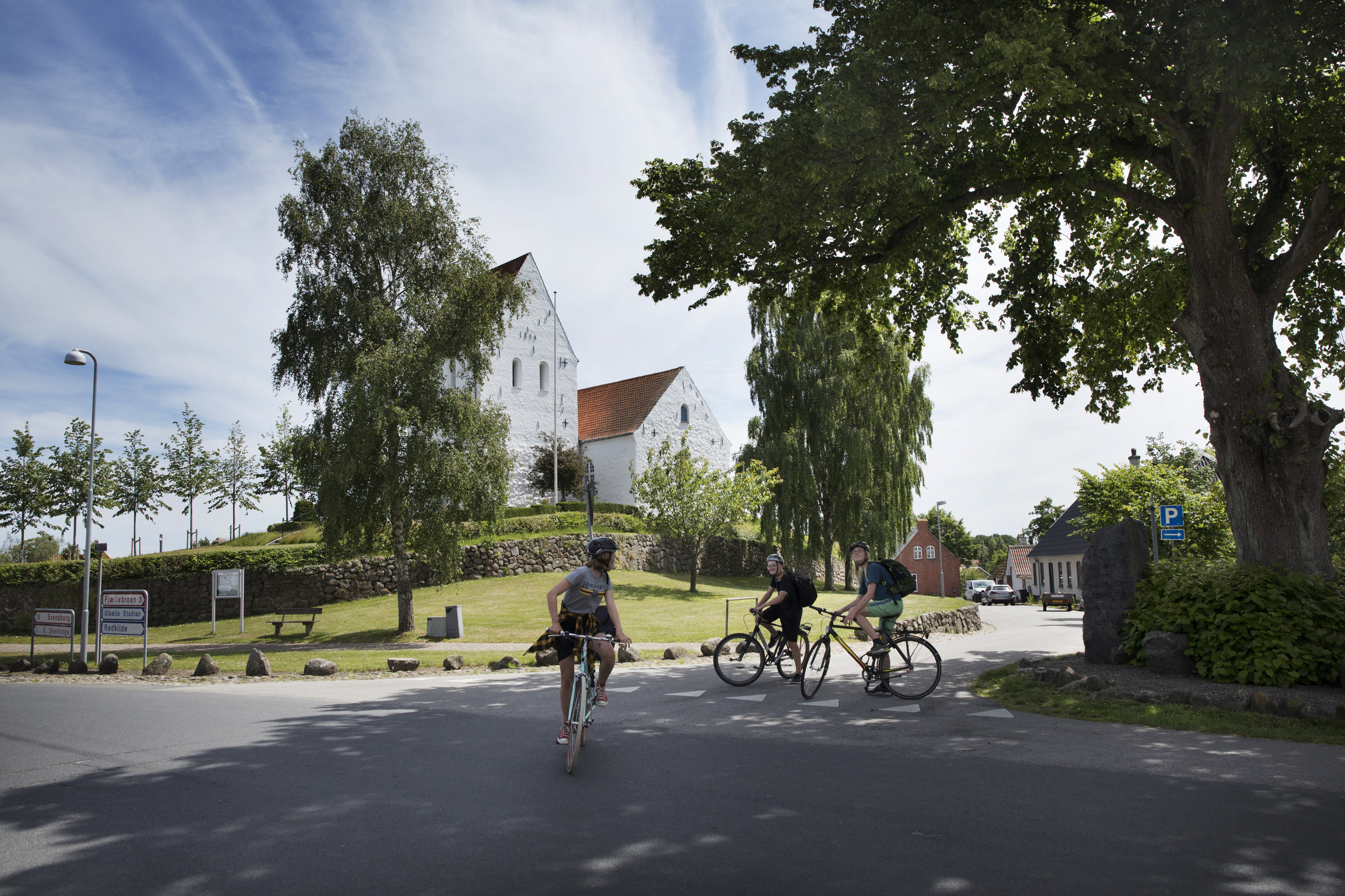 tre cykelister foran hvid kirke i grønne omgivelser